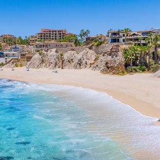 Pristine beach with turquoise waters and golden sand, nestled against dramatic cliffs with luxury resorts and palm trees dotting the coastline.