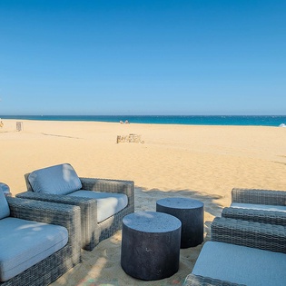 Private beachfront lounge featuring wicker seating with plush blue cushions set on golden sand, where turquoise waves meet endless blue skies and swaying palms frame your oceanfront escape.