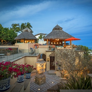 Stunning tropical resort architecture features thatched-roof palapas and stone terraces surrounded by lush palm trees and vibrant gardens.