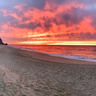 Dramatic sunset paints the sky in brilliant orange and pink hues over a pristine sandy beach with rocky cliffs.