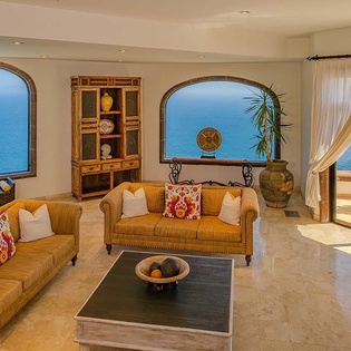 Picture yourself relaxing in this sunlit living space, where expansive arched windows frame breathtaking ocean vistas and gentle breezes drift through the open terrace doors. The warm tones and plush seating create your perfect seaside sanctuary.