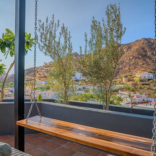 A hillside village with white-washed buildings cascades down the mountainside, visible beyond a rooftop terrace featuring a suspended wooden swing and planted greenery.