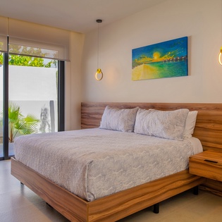 Wake up in your serene bedroom featuring a stunning wood platform bed, tropical garden views through floor-to-ceiling windows, and calming coastal artwork.