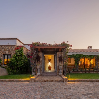 Elegant stone villa with warm lighting welcomes guests at twilight, featuring manicured gardens and traditional architecture.