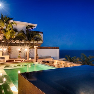 Luxurious beachfront villa featuring illuminated infinity pools, palm trees, and modern architecture against an evening ocean horizon under dramatic twilight skies.