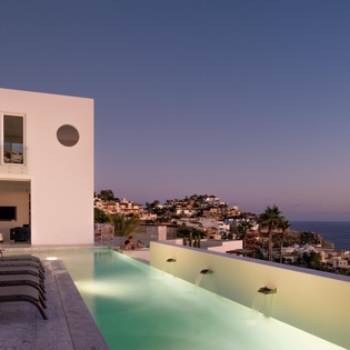 Modern villa with infinity pool overlooking coastal hillside town at twilight.