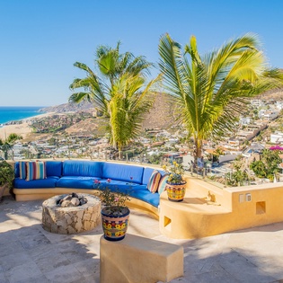 Stunning rooftop terrace showcases panoramic coastal views with vibrant blue seating, palm trees, and charming hillside neighborhood below.