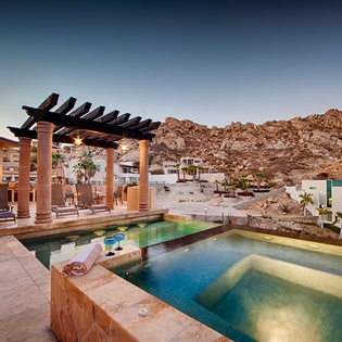 A dramatic hillside property with a multi-level pool and pergola-covered terrace, set against rugged desert mountains at dusk.