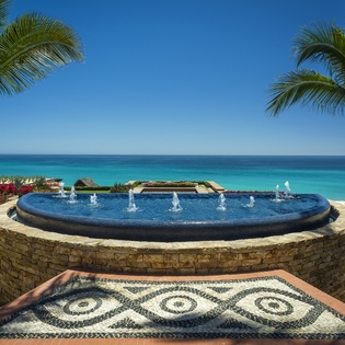 A stunning circular fountain with decorative mosaic detailing overlooks pristine turquoise waters and tropical palm trees.