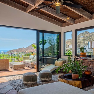 Step into your tranquil living room where rustic beams frame panoramic mountain and ocean views. Watch sunsets from the terrace, relax among lush greenery, and enjoy breezes through open glass doors.