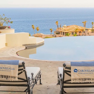 The property offers a stunning infinity pool overlooking the blue ocean, with lounge chairs perfectly positioned for your relaxation. The setting blends coastal luxury with tropical warmth, framed by surrounding villas and palm trees.