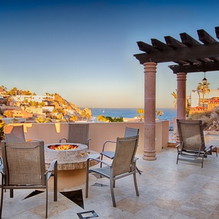 This hillside coastal property features an outdoor terrace with a fire pit, pergola, and expansive ocean views, framed by warm sunset lighting and surrounding neighborhood architecture.