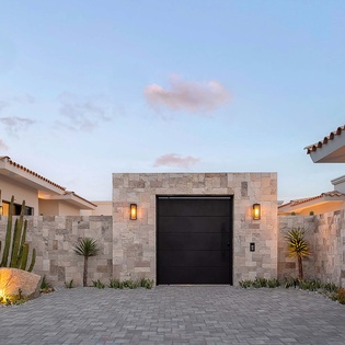 Elegant stone-clad entrance with modern desert landscaping welcomes visitors at dusk, featuring warm lighting and a paved courtyard.