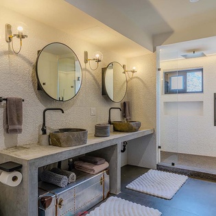 Step into your spacious bathroom with dual stone sinks, warm sconce lighting, and a charming vintage trunk for extra storage. The glass-enclosed shower and bright natural light create your perfect morning retreat.