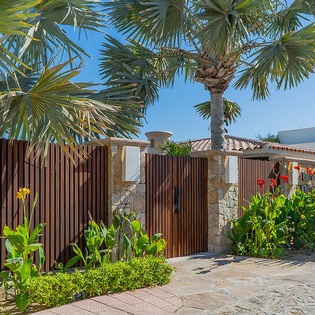 Private entrance gate set against a backdrop of tropical palms, leading to a sun-drenched Mediterranean-style villa.