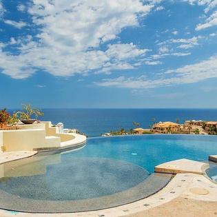 An expansive infinity pool overlooks the coastline, where sparkling blue water meets clear skies and neighboring oceanfront properties dot the landscape.