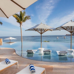 An infinity pool overlooking the ocean, featuring in-water lounge chairs under white umbrellas. Relax poolside with tropical palms swaying and coastal views stretching to the horizon.