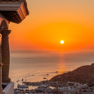 A stunning sunset view over the Mediterranean coastline, with traditional architecture framing the golden horizon and peaceful harbor below.