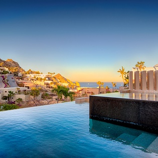An infinity pool overlooking a hillside coastal community during golden hour. The property sits among terraced homes carved into the rocky landscape, with ocean views stretching to the horizon.