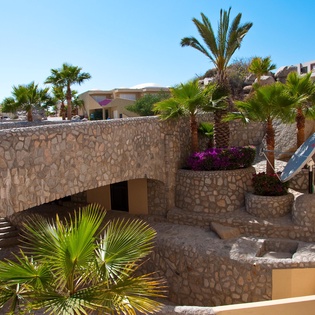 A sun-drenched property featuring terraced stone architecture, lush palm gardens, and vibrant bougainvillea blooms under clear blue skies.