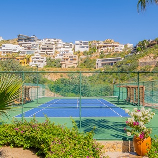 Professional tennis court surrounded by lush tropical landscaping and hillside residences in a scenic resort setting.