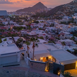 Aerial view of a coastal hillside community at dusk, with mountains rising dramatically in the background and the property nestled among palm trees and neighboring homes in a scenic seaside setting.