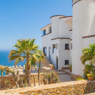 Stunning white-washed villa with distinctive turret architecture, lush tropical gardens, and blue chaise lounges overlooking the sparkling sea—a picturesque Mediterranean-style retreat.