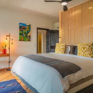 Your stylish bedroom features a plush bed with vibrant accent pillows, complemented by modern design touches like a sleek ceiling fan, warm lighting, and colorful artwork creating a welcoming retreat.