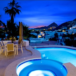 Stunning hillside villa overlooking illuminated Mediterranean village at twilight, featuring terrace pool and hot tub surrounded by palm trees.