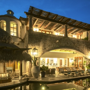 Stunning beachfront villa illuminated at twilight, featuring traditional palapa roofing and stone archways surrounding a peaceful reflecting pool.