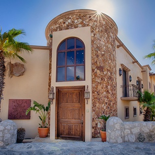 Mediterranean-style villa with distinctive stone turret entrance, arched wooden door, and palm-lined courtyard in a hillside setting.