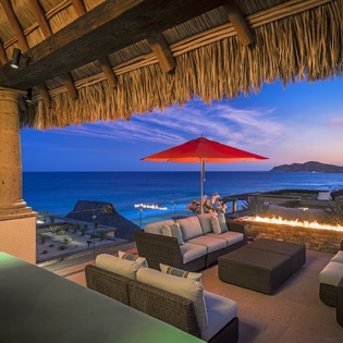 Spectacular oceanfront terrace with traditional thatched roof, elegant seating, fire feature, and dramatic sunset views over the coastline.