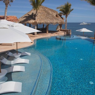 A luxury infinity pool meets pristine turquoise waters, where palm-shaded loungers invite you to relax under endless blue skies and gentle ocean breezes.