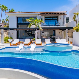 Modern luxury villa with curved swimming pool and hot tub, surrounded by tropical palms in a sunny resort setting.