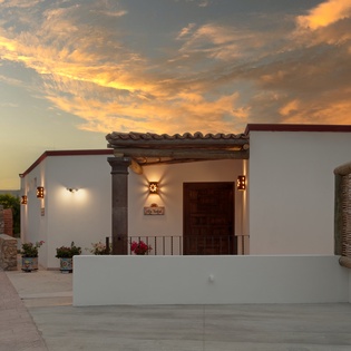 A charming villa entrance welcomes guests beneath a dramatic golden sunset sky, showcasing Mediterranean architecture and warm evening lighting.