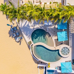 Aerial view showcasing a uniquely curved pool surrounded by lush palm trees and beachfront access, where the property meets golden sands and turquoise waters.