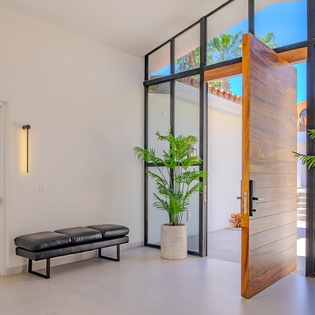 Step into your stylish entrance where modern design meets tropical warmth. Floor-to-ceiling glass doors open to reveal lush palms and blue skies awaiting you.