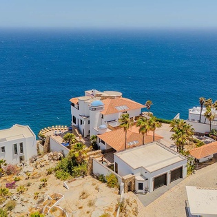 Coastal properties with terracotta roofs, palm trees, and stunning ocean backdrop create a picturesque Mediterranean-style seaside neighborhood in this aerial view.