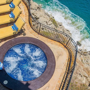 Aerial view of the property's dramatic oceanfront location, featuring a circular hot tub, yellow sun loungers on a curved terrace, and direct access to the rocky coastline and turquoise waters below.