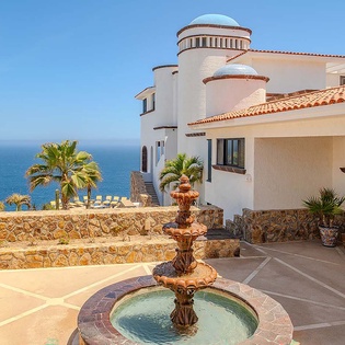 Mediterranean-style villa with white stucco walls, terracotta tile roof, turret, stone courtyard fountain, and dramatic ocean views set against clear blue skies.