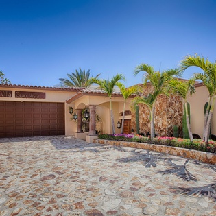 Villa Gran Vista welcomes guests with its warm stucco facade, lush tropical landscaping, vibrant blooms, and Spanish-tile accents under clear blue skies.