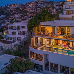 Hillside luxury villa with illuminated terraces and infinity pool at twilight, overlooking the neighboring coastal community and surrounding landscape.