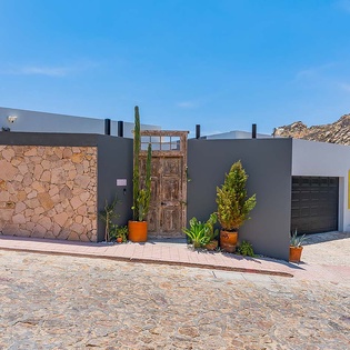 Modern property entrance features natural stone accents and desert landscaping, nestled against dramatic mountain backdrop. Cobblestone driveway and rustic wooden door create a welcoming arrival experience.