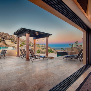 Stunning sunset views frame this spacious terrace featuring a shaded pergola, lounge chairs, and infinity pool, all set against dramatic mountain and ocean scenery.