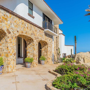 A stunning villa entrance featuring warm stone archways, vibrant desert landscaping with flowering plants, and sweeping coastal views under clear blue skies.