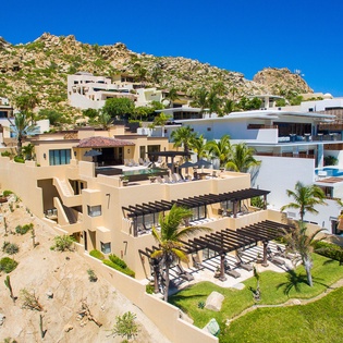 Hillside resort community nestled against dramatic rocky peaks, featuring modern villas with private terraces, lush gardens, and palm trees under clear blue skies.
