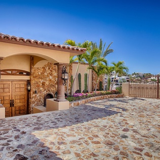 A Spanish-colonial estate features terracotta tile roofing, cobblestone driveway, stone archway entrance, tropical landscaping, and an ornamental gate overlooking hillside homes.