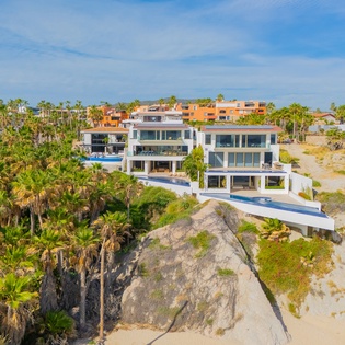 Modern beachfront villa perched on dramatic rocks with tropical palms and pristine sandy beach access.