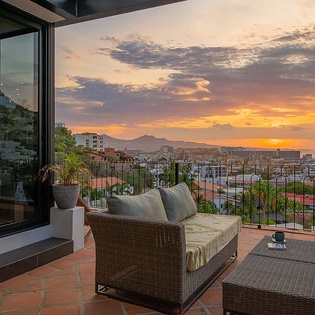 A private terrace frames a breathtaking sunset over mountains and cityscape. The wicker lounge invites evening relaxation as the sky transforms into warm, vibrant hues.
