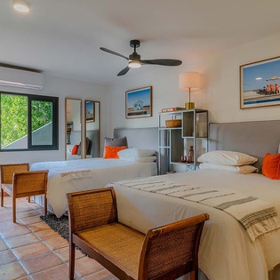 Your bright, airy bedroom features two comfortable beds with crisp linens and pops of orange accents. Natural light pours through large windows overlooking lush greenery, while modern amenities keep you cool and relaxed.